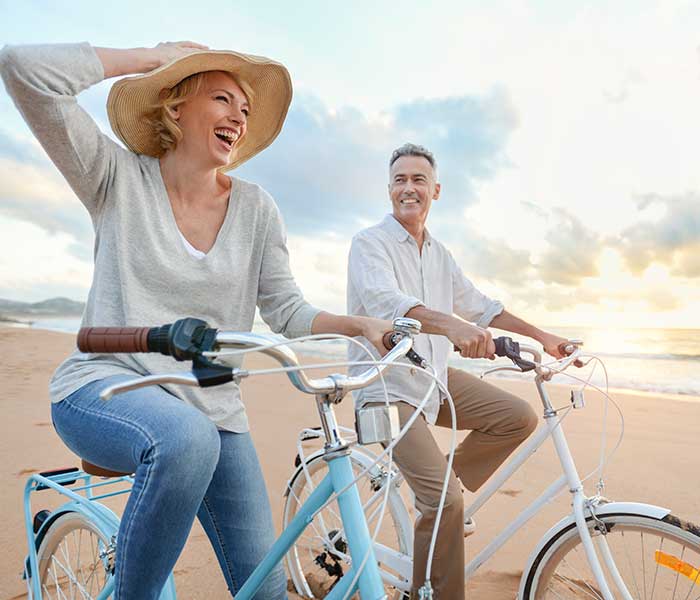 Mature Couple Cycling on the Beach Highlighting Recent Dentures at S&L Dental
